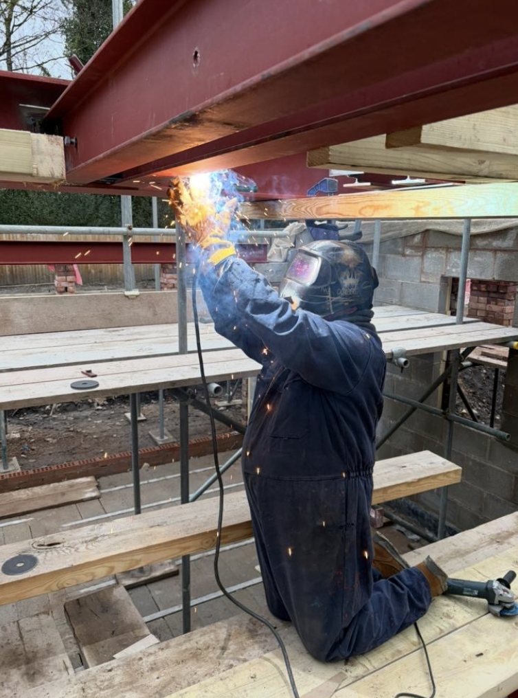 Welder joining red structural steelwork overhead with sparks flying